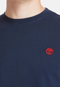 Mörkblå bomullströja med rund hals, med en liten röd broderad trälogo på vänster bröstområde. Slät textur.