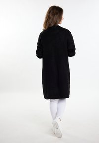 Cardigan lung de tricotaj negru, cu un model texturat, prezentând un guler înalt și mâneci supradimensionate. Purtați peste colanți albi și adidași.