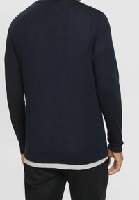 Maglione a maniche lunghe blu navy realizzato in morbido tessuto a maglia, con colletto tondo e polsini a costine, con una camicia bianca sovrapposta visibile sotto.