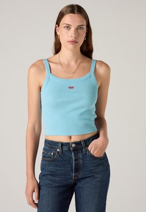 Jeune femme aux longs cheveux bruns porte un débardeur bleu clair et un jean bleu foncé, debout avec une main dans la poche devant un fond uni.