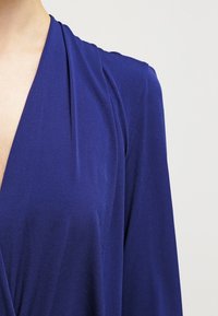 Vestido cruzado azul real con un profundo escote en V, mangas largas y una tela suave y lisa. Presenta un sutil detalle de drapeado a lo largo del escote.
