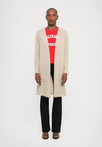 Beige gestrickte Strickjacke mit strukturiertem Muster, offenen Vorderseite und Fransenbesatz, über einem roten Grafik-T-Shirt und schwarzen Hosen getragen.