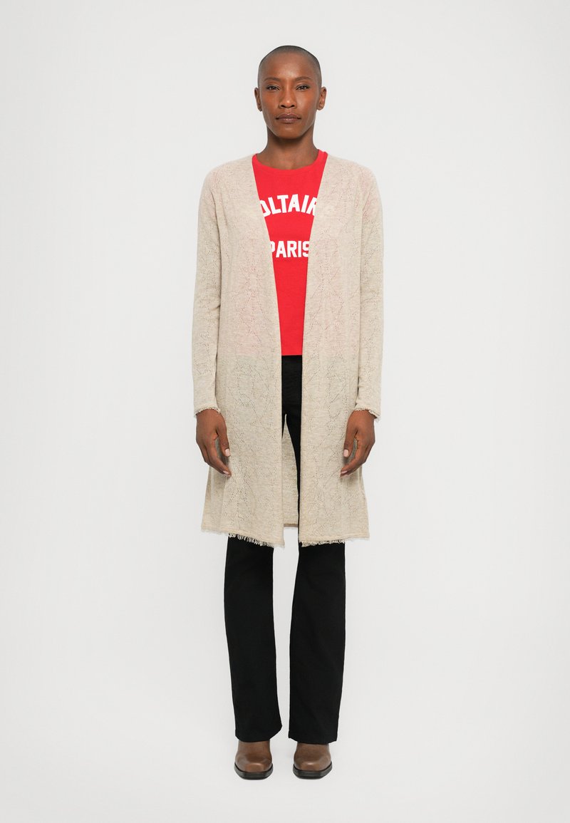 Beige knitted cardigan with a textured pattern, open front, and fringe hem, layered over a red graphic t-shirt and black pants.
