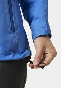 Niebieska wodoodporna kurtka z zamykaną na zamek kieszenią i regulowanym sznurkiem u dołu. Zbliżenie na rękę chwytającą sznurek.