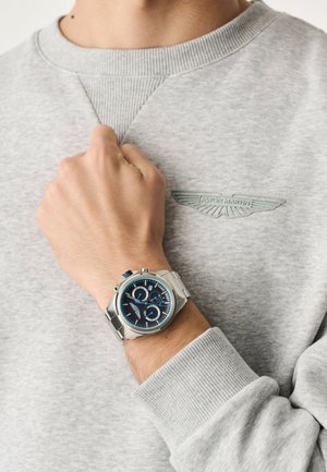 Ceas cronograf argintiu cu cadran albastru, marcaje albe pentru ore și un accent albastru. Purtați pe încheietura mâinii peste un hanorac gri deschis.