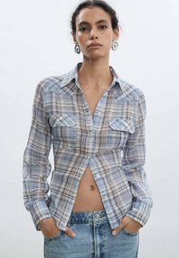 Blue and white plaid shirt with long sleeves, two chest pockets, and a button-down front; paired with high-waisted denim shorts.