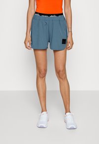 Ljusblå idrottsshorts med ett elastiskt svart midjeband med logotyp, sidofickor och en diskret glansig yta, tillsammans med vita sneakers.