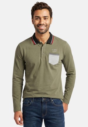 Homme souriant portant un polo manches longues vert olive avec un col noir et une poche poitrine grise, associé à un jean bleu, debout sur fond blanc.