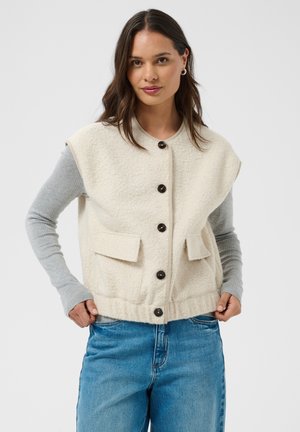 Femme aux longs cheveux foncés portant un gilet beige boutonné sur un haut gris à manches longues et un jean bleu, debout devant un fond uni.