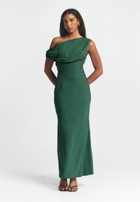 Dlouhé zelené šaty s jedním ramínkem, s drapériemi a úzkým střihem. Mají hladkou texturu a splývavou siluetu.