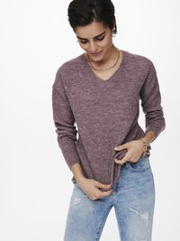 V-Ausschnitt Pullover aus strukturiertem maulbeerenfarbenem Wollmix, getragen mit hellblauen, verwaschenen Jeans, die Hände am Saum positioniert. Goldene Kettenhalskette sichtbar.