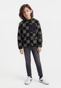 Barn med skulderlangt brunt hår iført sort og gråternet fleecejakke, mørke jeans og hvide sneakers, stående mod en hvid baggrund.