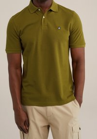 Olivgrön polo med texturerad tyg, klassisk krage och ett litet broderat emblem på vänster bröst.