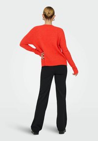 Femme aux cheveux blonds attachés en chignon, portant des boucles d'oreilles en or, un pull orange, un pantalon noir et des chaussures noires, posant devant un fond blanc.