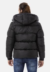 Schwarze Daunenjacke mit glänzender Kapuze, gestepptem Design und elastischen Bündchen. Kombiniert mit blauen Jeans, was ein lässiges Aussehen unterstreicht.