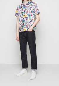 Camisa de manga corta colorida con varios patrones, combinada con jeans de mezclilla oscuros. Zapatillas blancas completan el look. Estilo y ajuste casual.