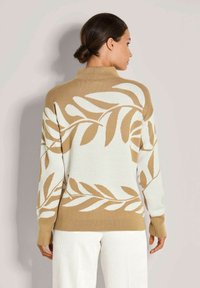 Frau mit dunklem Haar zu einem Dutt frisiert, trägt einen beigen und weißen Pullover mit großem Blattmuster, kombiniert mit weißen Hosen, mit dem Rücken zur Kamera.