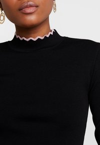 Černé žebrované tričko s rolákem a vlnitou růžovou lemovkou u výstřihu. Hladká textura s přiléhavým střihem. Jsou viditelné zlaté kruhové náušnice.