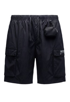 Junge Frau mit nassen Haaren, die ein weißes ärmelloses Top und schwarze Cargo-Shorts mit Kordelzug trägt, an denen eine kleine Tasche befestigt ist.