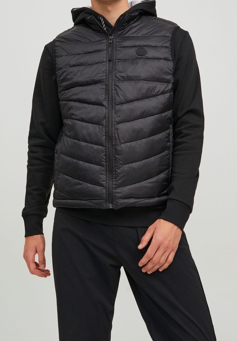 Gilet imbottito nero con chiusura a zip, caratterizzato da pannelli trapuntati orizzontali, cappuccio e patch del logo sul petto, indossato sopra una maglietta nera a maniche lunghe.