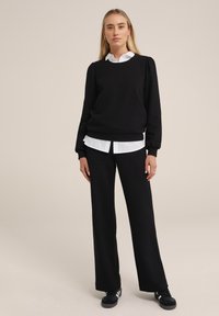 Hanorac negru tricotat cu guler rotund, mâneci lungi și manșete din bumbac, asortat cu o cămașă albă cu guler și pantaloni negri cu croială largă.