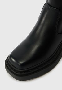 Bottine en cuir noir avec un bout carré et une semelle en caoutchouc chunky sur fond blanc.