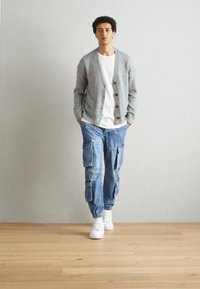 Cardigan gris claro con botones, camiseta blanca, pantalones cargo azules con bolsillos y zapatillas blancas sobre un suelo de madera contra una pared neutra.