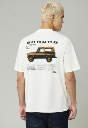 T-shirt en coton blanc à manches courtes, arborant un graphique rétro d'un véhicule Bronco et un texte au dos en impression noire et brune.