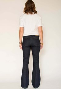 Jeans évasés bleu marine à taille haute, fabriqués en denim. Comprend des poches arrière et des détails de couture dorée. Portés avec un t-shirt blanc.