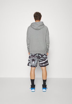 Mann trägt grauen Kapuzenpullover, schwarze und graue Raiders-Logo-Shorts, schwarze Socken und blau-weiße Turnschuhe, steht vor einer weißen Wand.
