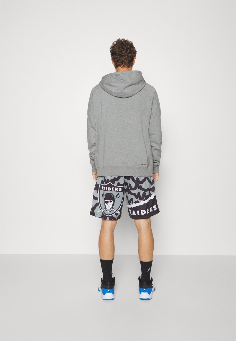 Homme portant un sweat à capuche gris, un short noir et gris avec le logo des Raiders, des chaussettes noires et des baskets bleues et blanches, face à un mur blanc.