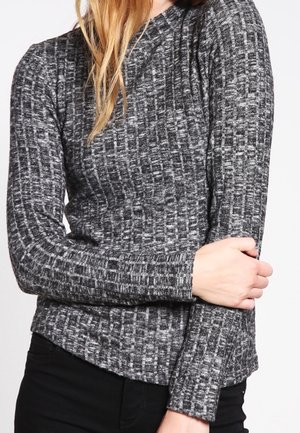 Personne portant un pull texturé gris et noir à manches longues, un bras croisé tenant l'avant-bras opposé.