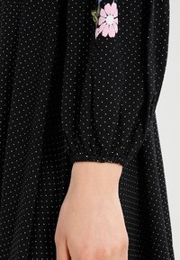 Vestido negro con lunares blancos, con mangas abullonadas elásticas y bordado de flores en rosa en el lado izquierdo. Tela suave y ligera.