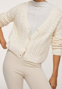 Kobieta w kremowej dzierganej kardiganie z perłowymi zdobieniami nosi biały top oraz beżowe spodnie z wysokim stanem.