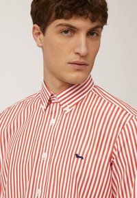 Camisa de botones a rayas rojas y blancas con cuello, que presenta un pequeño logo azul en el lado izquierdo. La textura de la tela parece suave.