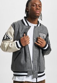 Grå ullblandings varsityjakke med kremfargede skinnermedaler, stripete krage og mansjetter, stort bokstavpatch og intrikate detaljer på ermene.