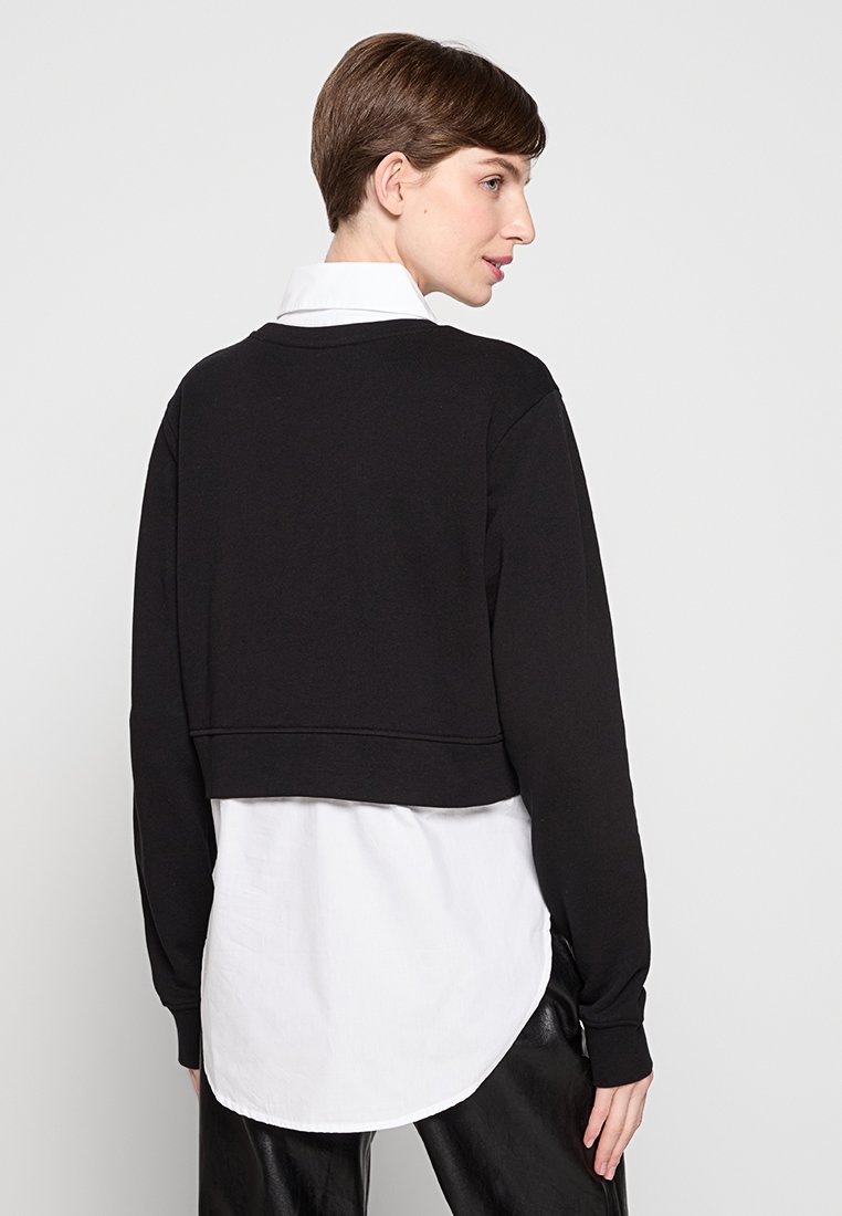 Sweatshirt noir coupé avec manches longues, col rond, tissu texturé, superposé sur une chemise blanche à l'ourlet arrondi et texture lisse.