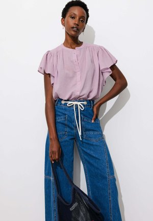 Femme portant un chemisier violet clair à manches volantées et un jean bleu taille haute, tenant un sac cabas en maille noire sur fond blanc.