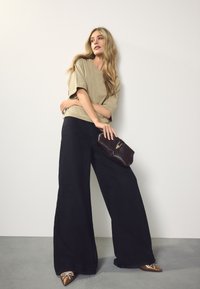 Beige oversized top, sorte bukser med vide ben, sort clutch med hardware-detalje og spidse brune pyntede sko. Glatte teksturer overalt.