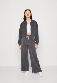 Grijze, gestructureerde velours trainingspak bestaande uit een cropped jasje en brede broekspijpen. Witte trekkoord op de broek; model draagt witte sneakers.