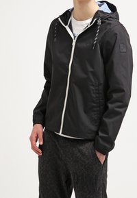 Personne portant une veste à capuche noire zippée avec des cordons blancs et un pantalon sombre à motifs, une main dans la poche.