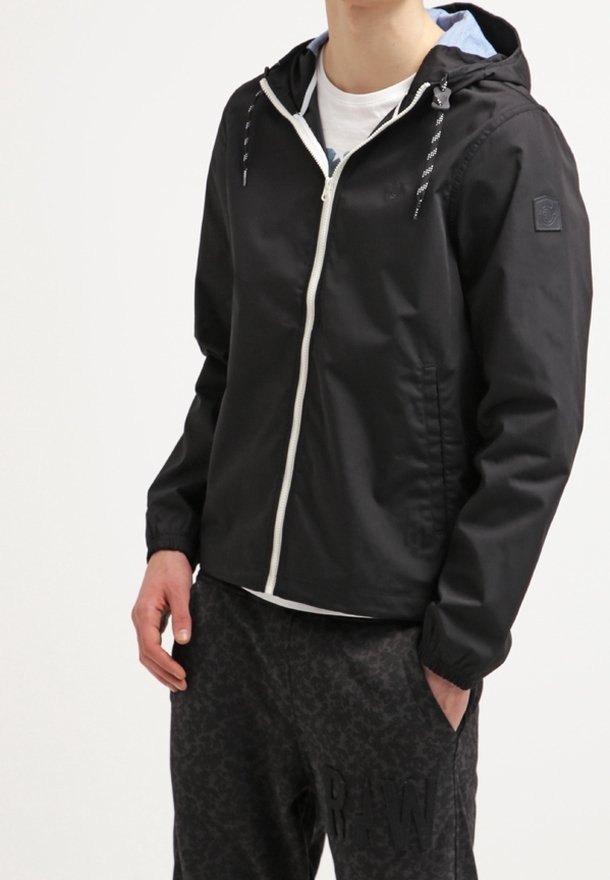 Personne portant une veste à capuche noire zippée avec des cordons blancs et un pantalon sombre à motifs, une main dans la poche.