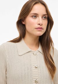 Sweter kardigan w jasnobeżowym kolorze, z teksturowanym wzorem, kołnierzykiem oraz pięcioma dużymi, błyszczącymi guzikami z przodu.