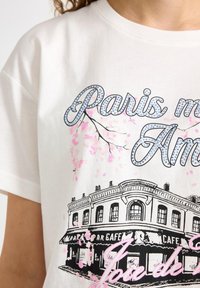 Hvid bomulds-T-shirt med sort caféillustration, lyserøde kirsebærblomstdetaljer og teksten "Paris mon Amour" i glitrende blå.