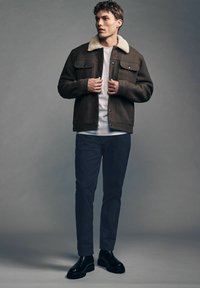 Chaqueta de lana marrón con un cuello de borrego blanco, llevada sobre una camisa blanca, combinada con pantalones oscuros y botas negras. Corte ajustado.
