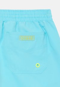 Costumi da bagno blu brillante con vita elasticizzata, tasca posteriore con etichetta gialla "GUESS" e bottone di accento verde.