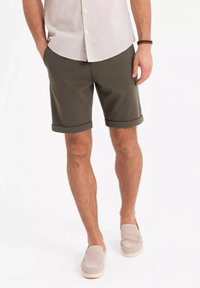 Gröna tygshorts med rullade fållar och en texturerad yta. Parat med en ljusfärgad skjorta med knappar och beigefärgade slip-on skor.