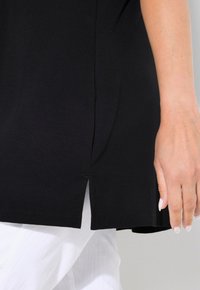 Černý top vyrobený z lehkého materiálu, s křivkami na spodním okraji a bočními rozparky, v kombinaci s bílými texturovanými kalhotami.