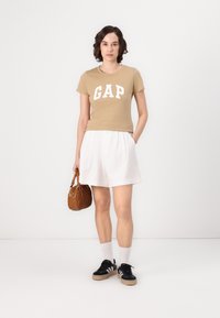 Light brown fitted t-shirt with "GAP" in white letters, paired with white pleated shorts, black sneakers with white accents, and a small brown handbag.