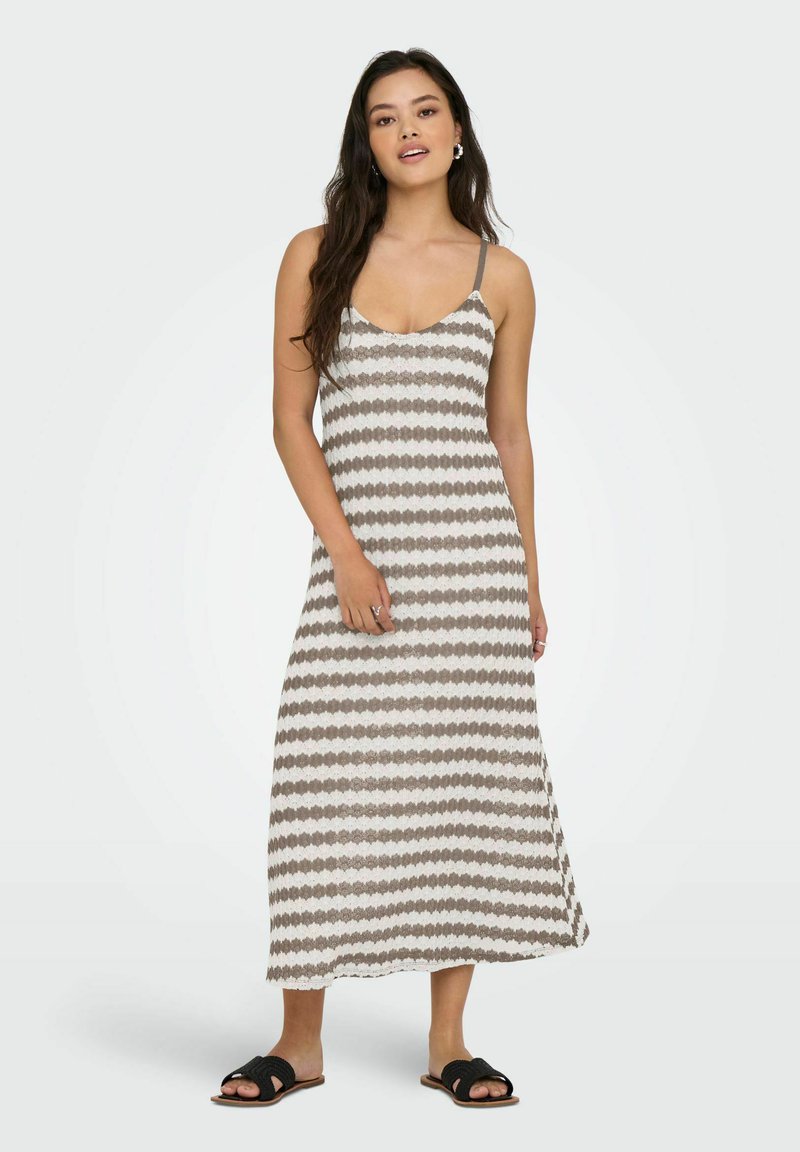 Femme aux longs cheveux foncés portant une robe maxi rayée sans manches et des sandales noires de type slide, debout sur un fond uni clair.